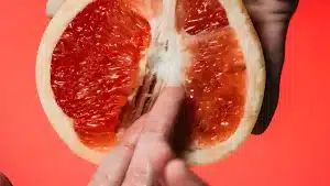 A close up of a hand touching the juicy red pulp of a halved grapefruit against a bright coral background