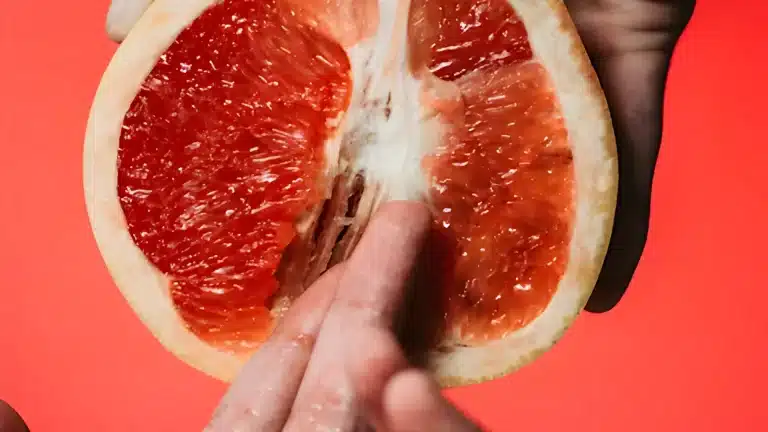 A close up of a hand touching the juicy red pulp of a halved grapefruit against a bright coral background