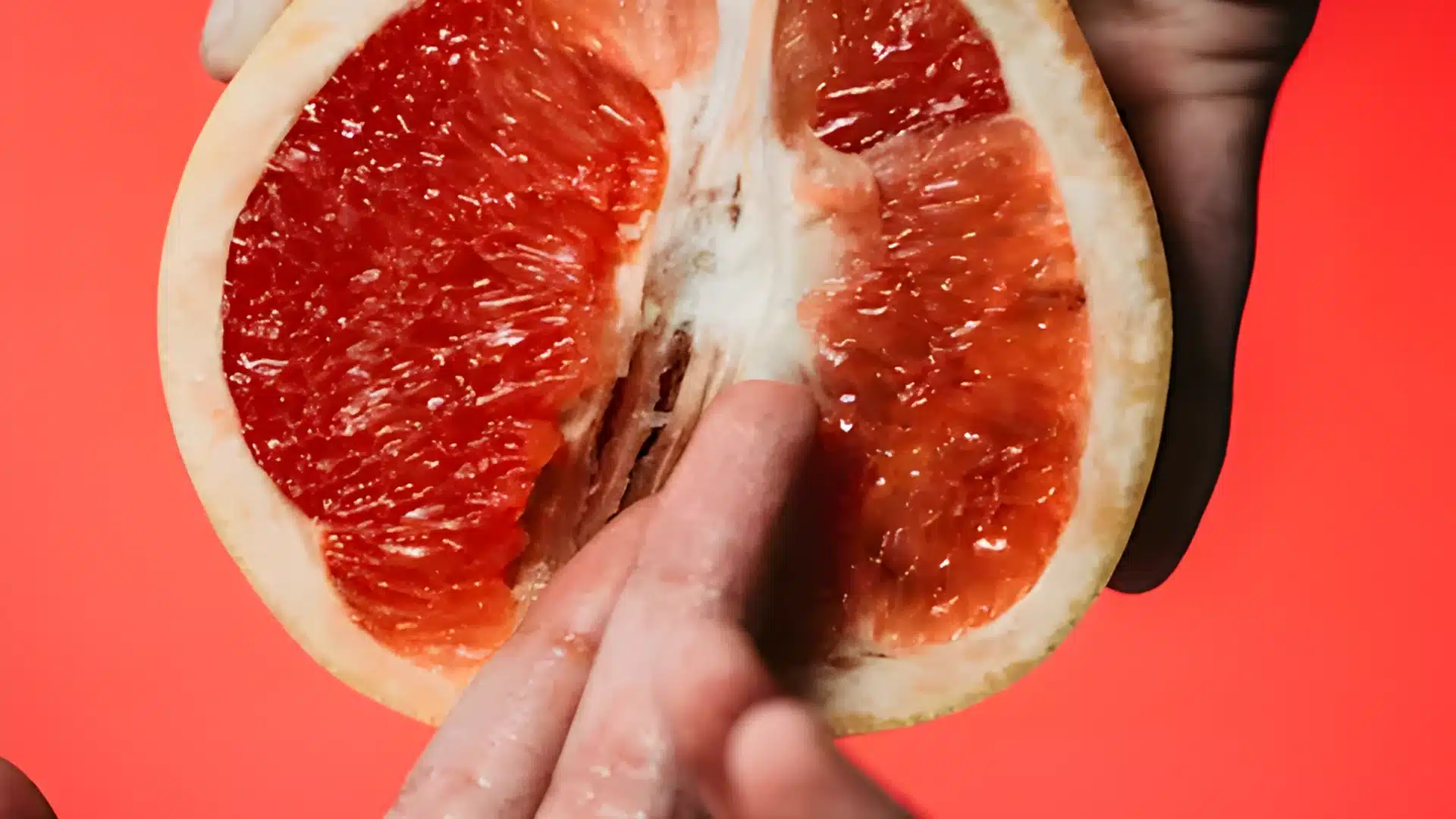 A close up of a hand touching the juicy red pulp of a halved grapefruit against a bright coral background