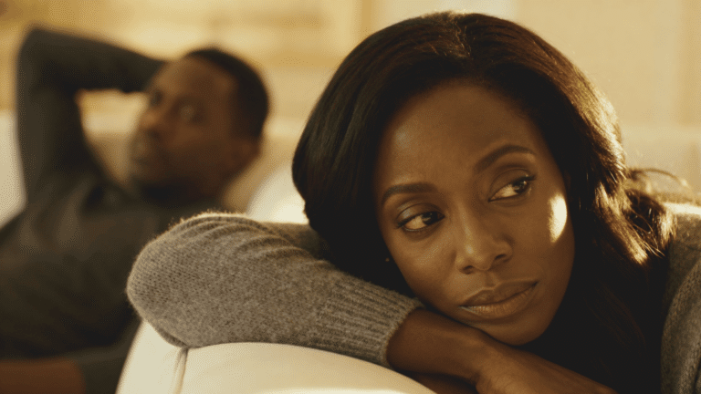 A contemplative woman rests her head on her arms while a blurred man sits in the background.