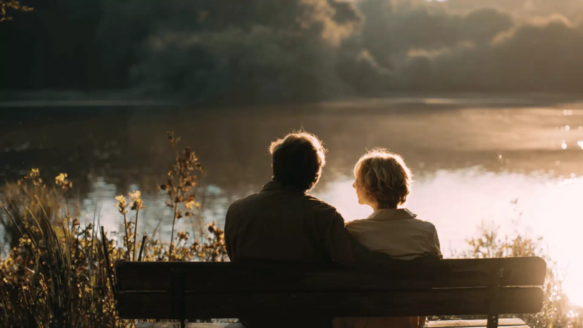 A couple sits on a wooden bench looking at a calm lake during a golden sunset with green trees in the soft background