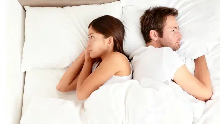 A man and a woman lie in bed back to back with upset faces showing a tense conflict between the couple in white linens
