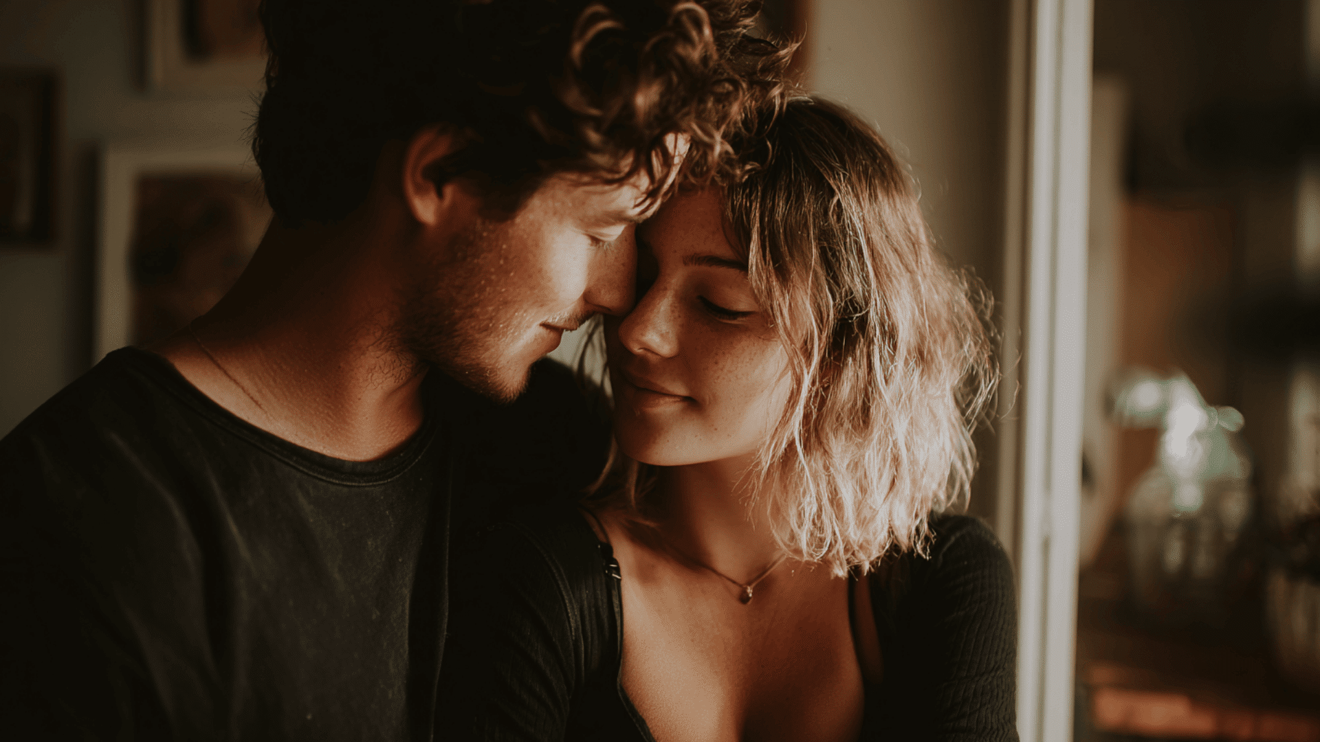 A man and woman sit indoors at a table in soft light while looking at each other with quiet and thoughtful expressions