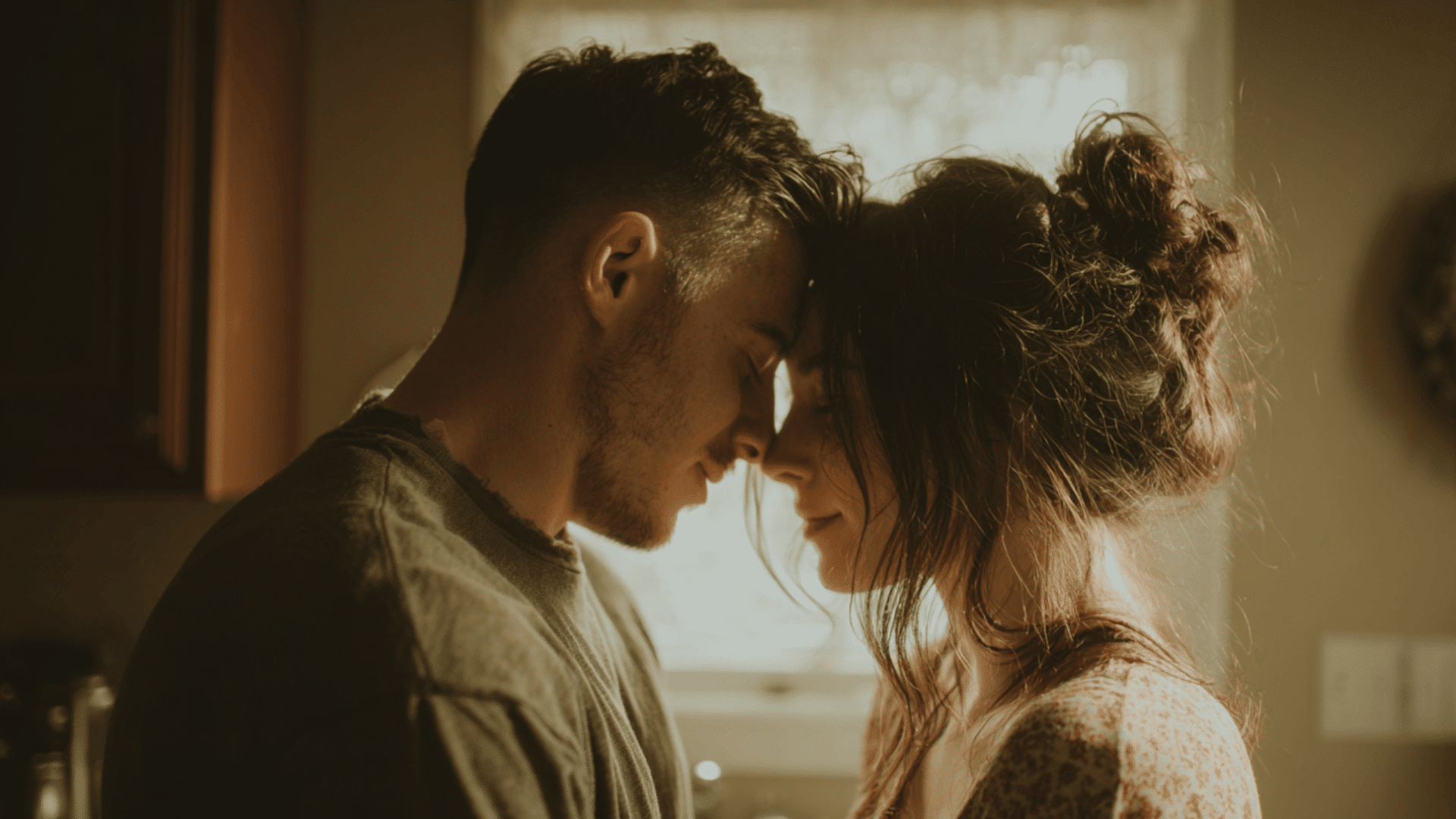 A man and woman stand close in a kitchen with foreheads touching in soft warm light as they share a quiet cozy moment