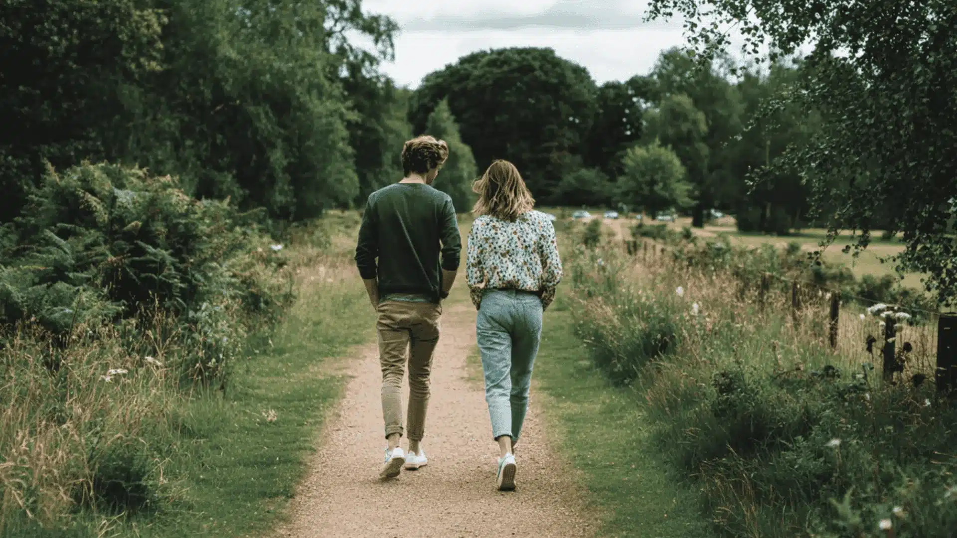A man and woman walk away down a dirt path through a green grassy field with trees under a soft cloudy sky in the summer (1)