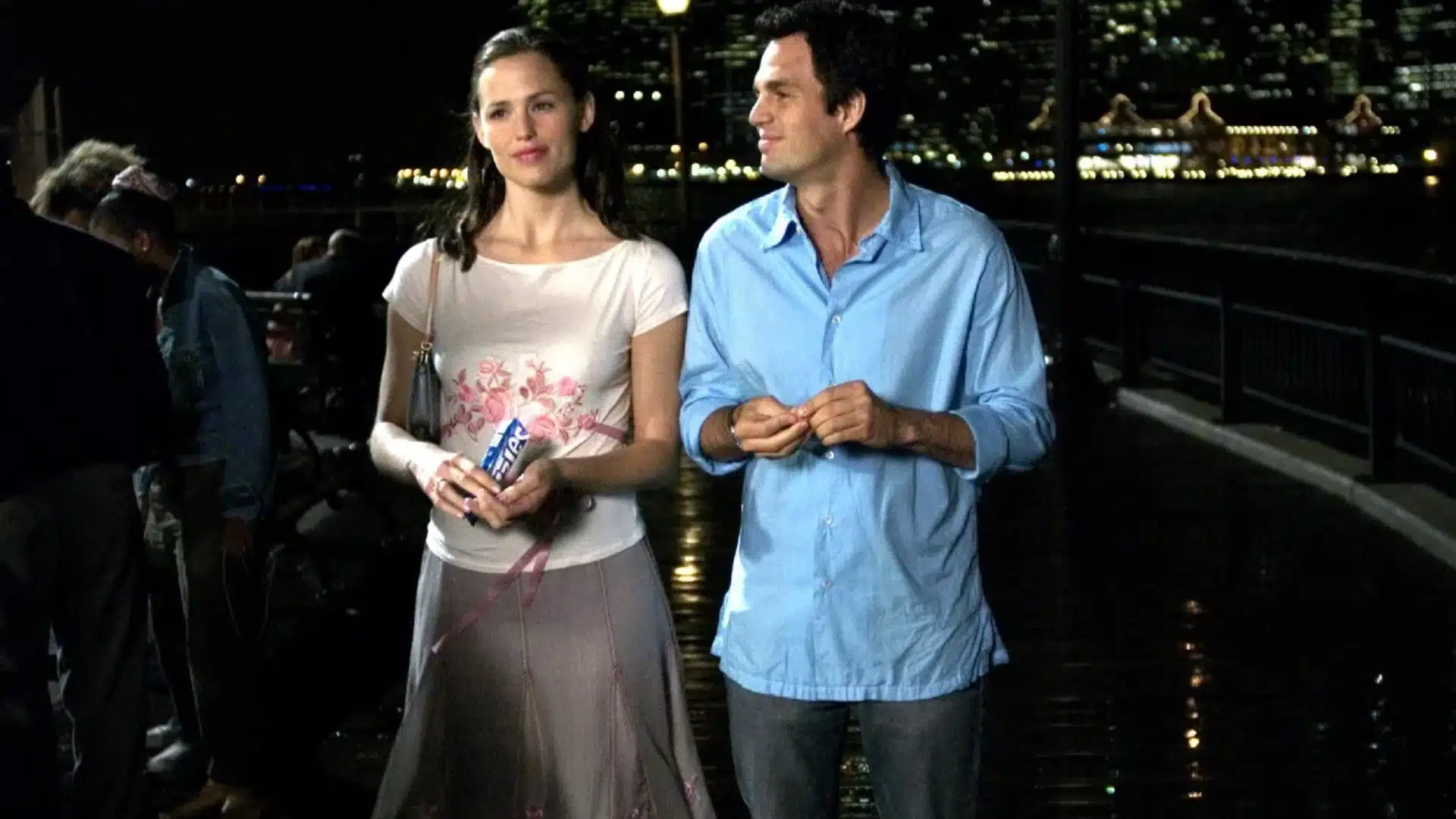 A man in a light blue shirt and a woman in a white floral top stand outdoors at night near water with city lights blurred in the background.