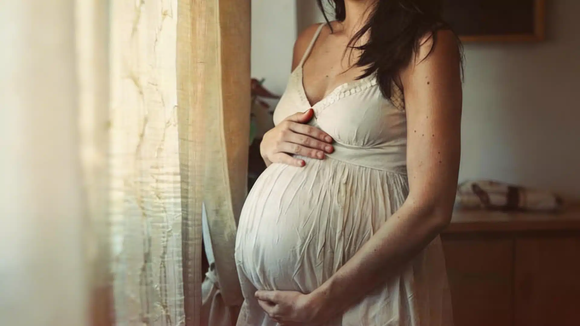 A pregnant woman in a white dress stands by a window holding her belly in a warm sunlit room with soft curtains and light