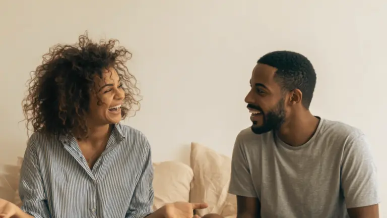 A smiling couple laughing together while seated indoors against a neutral background with beige pillows