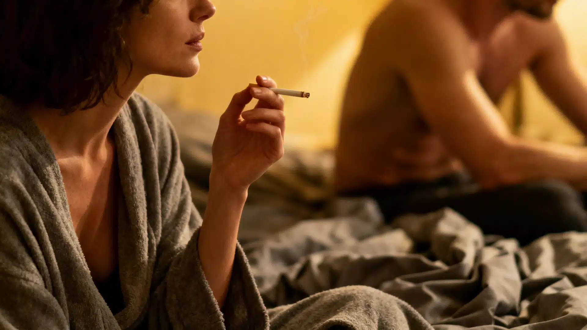 A woman in a grey robe smoking a cigarette on rumpled bed sheets with a shirtless man blurred in the background against a yellow wall