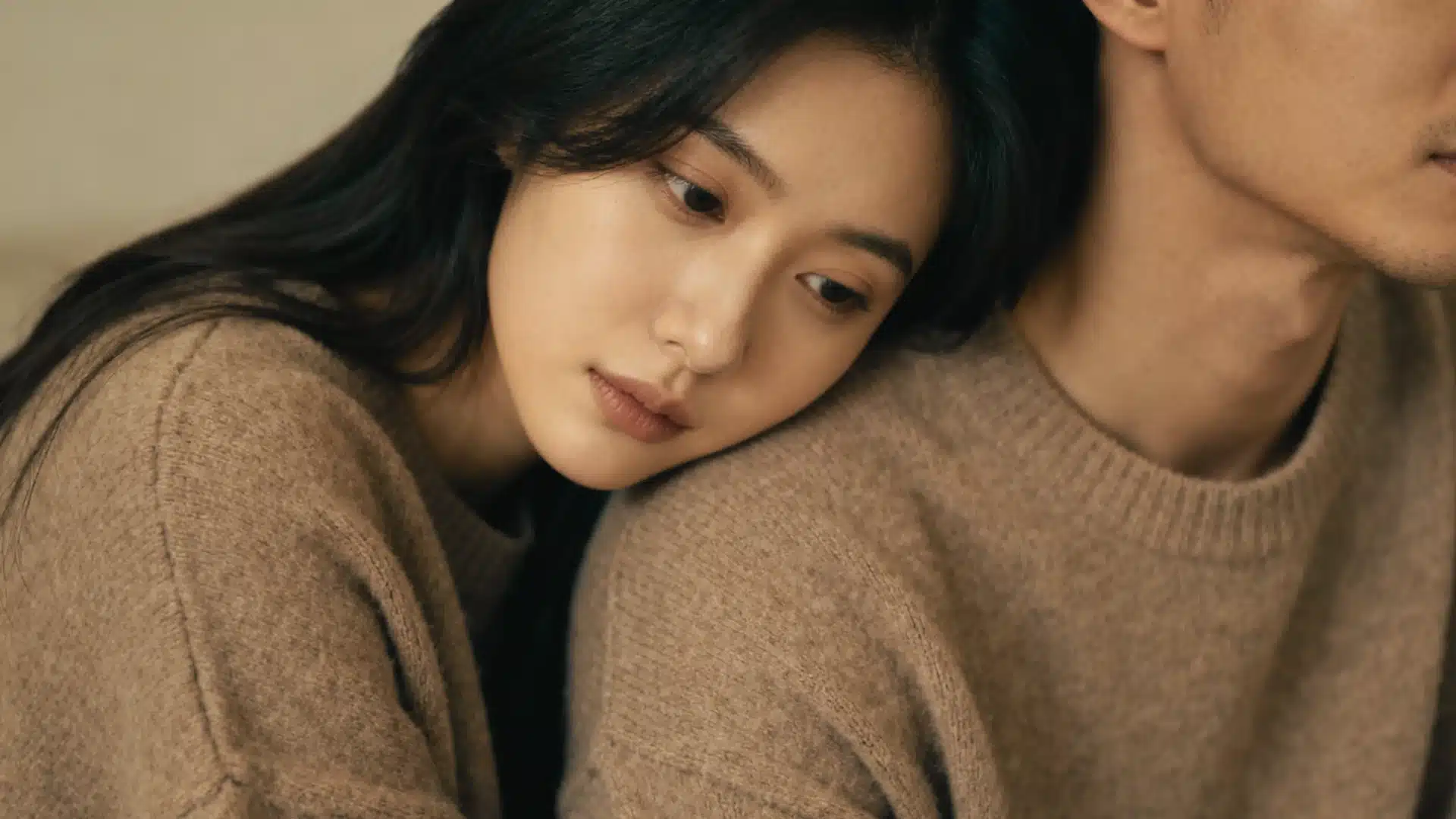 A woman with long dark hair leans her head gently on a man's shoulder, both wearing cozy beige sweaters