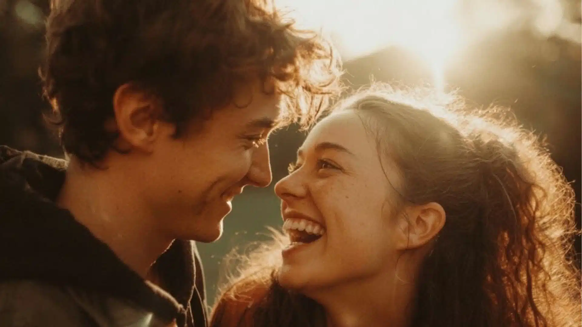 A young man and woman look lovingly at each other laughing warmly outdoors during the golden hour sunset light.