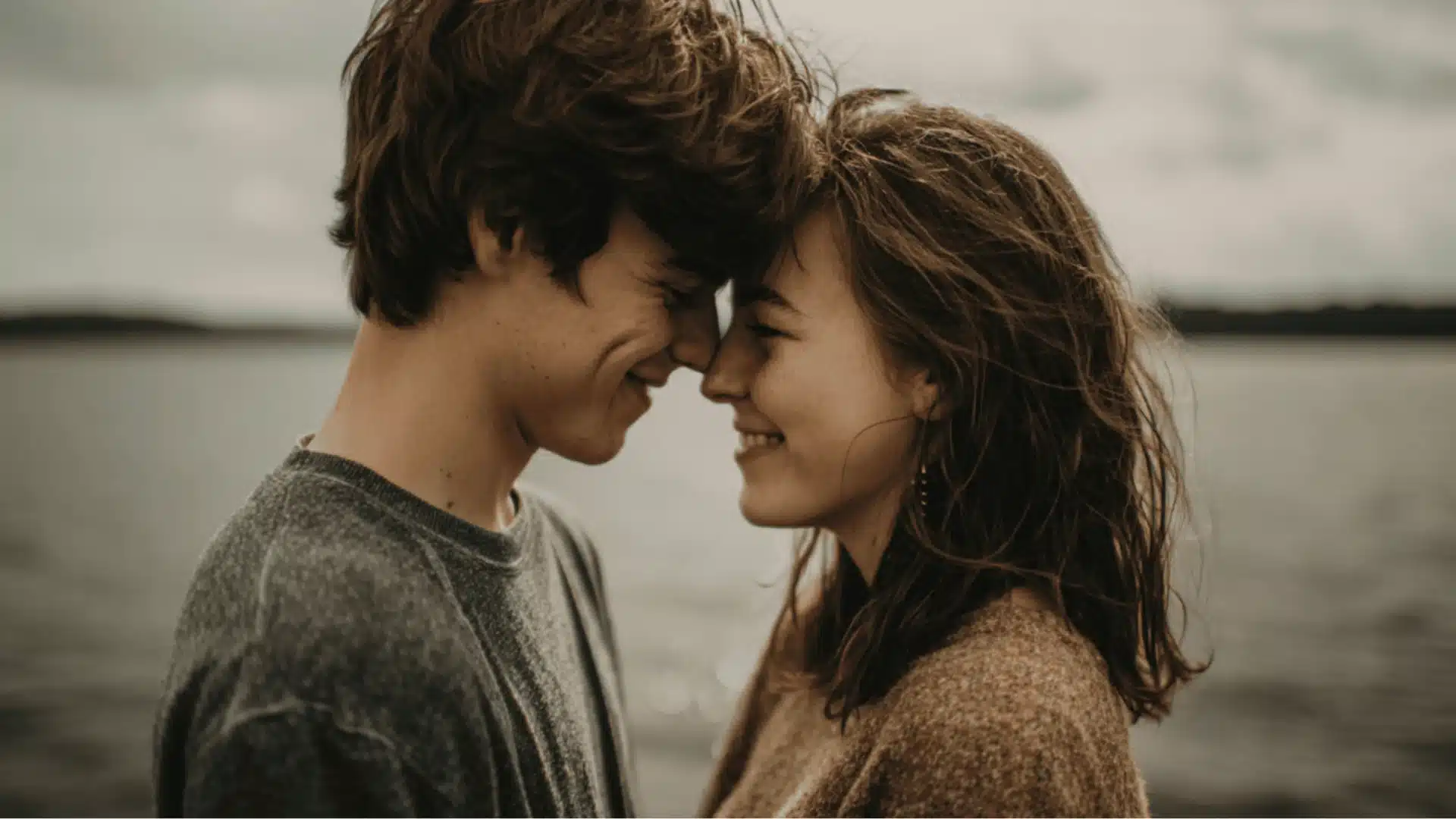 A young man and woman touch foreheads and smile together while standing by a lake under a gray sky on a quiet cloudy day (3)