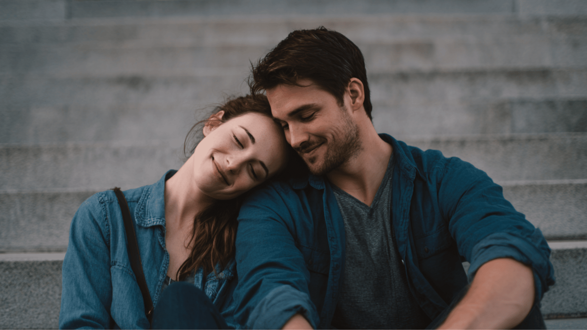 A young woman leans her head on a smiling mans shoulder while both sit together on wide stone steps outside a building