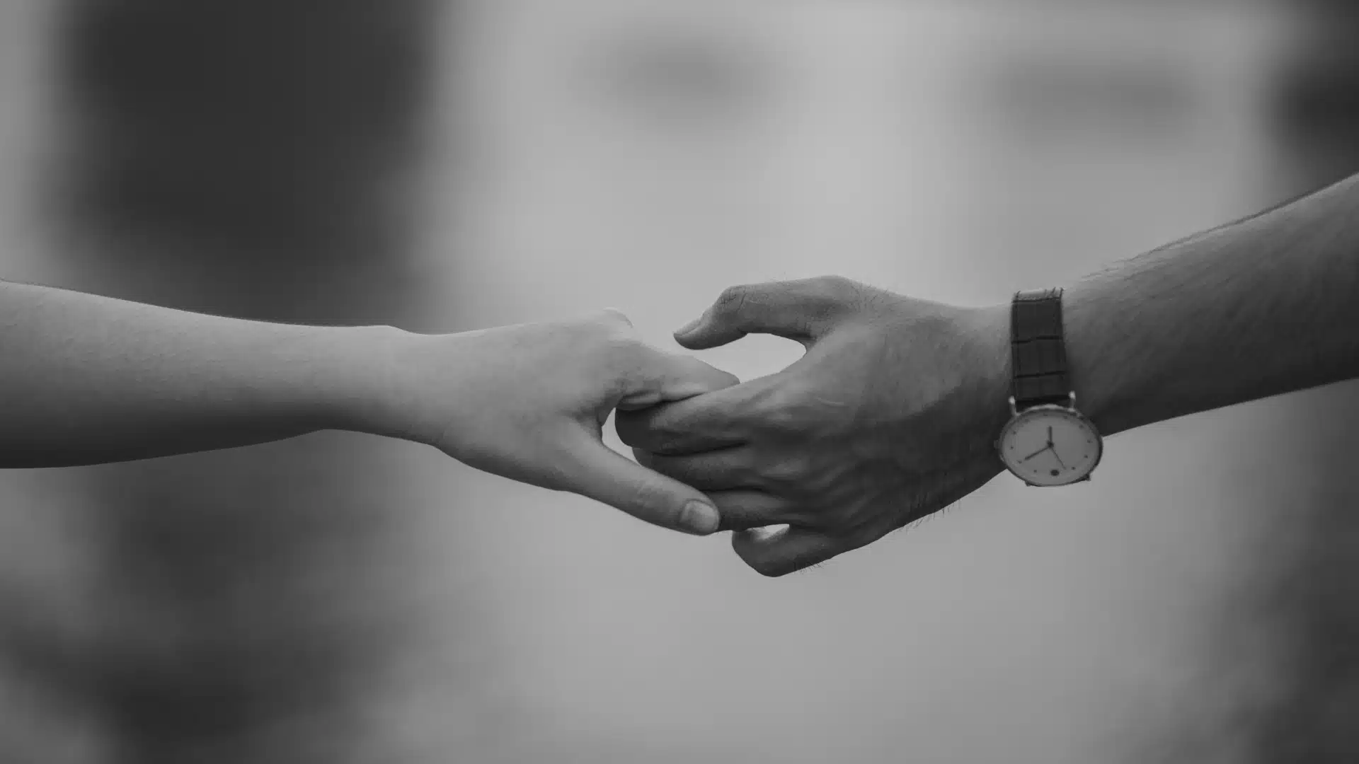 Black and white image of two hands gently touching, with one arm wearing a wristwatch, against a blurred background