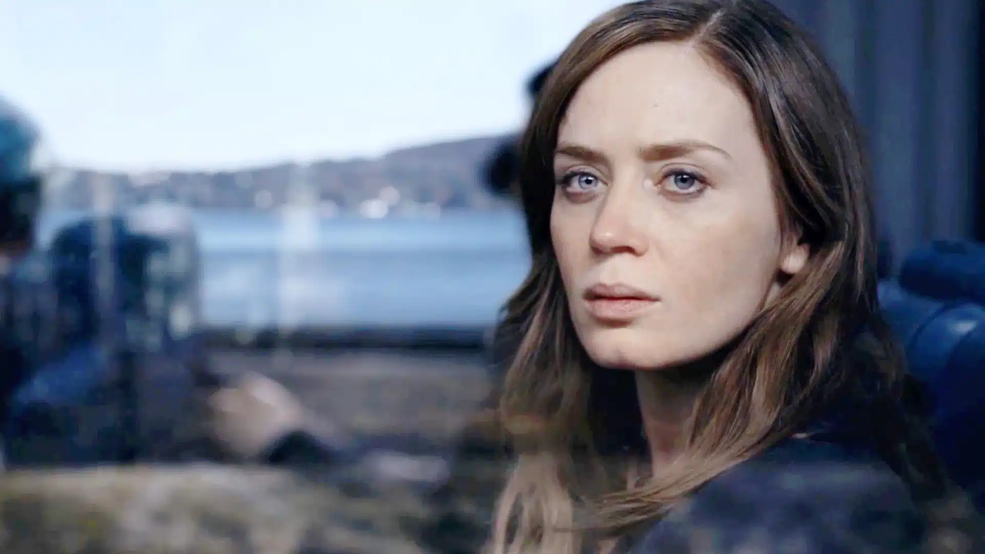 Close-up of Emily Blunt with intense blue eyes looking off-camera against a blurred background of water and distant land