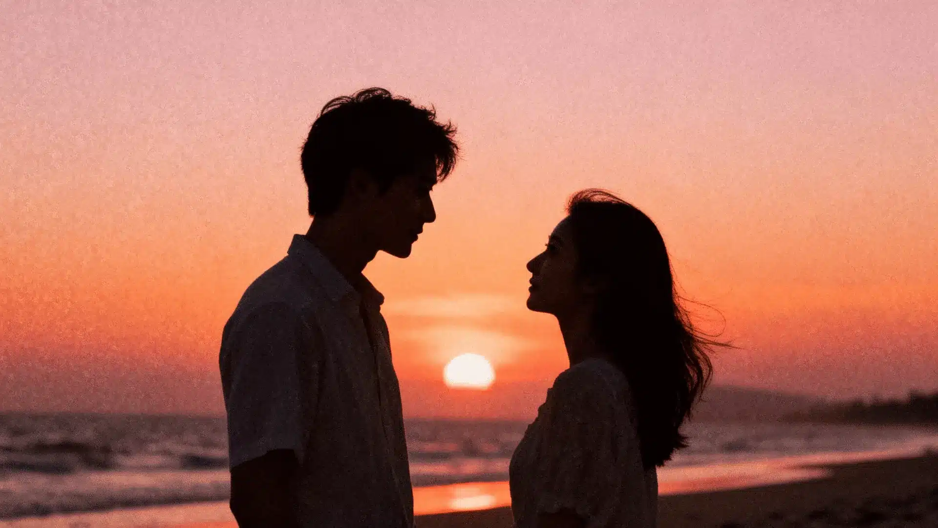 Couple silhouettes facing each other on a beach at sunset with warm orange sky and ocean waves in background