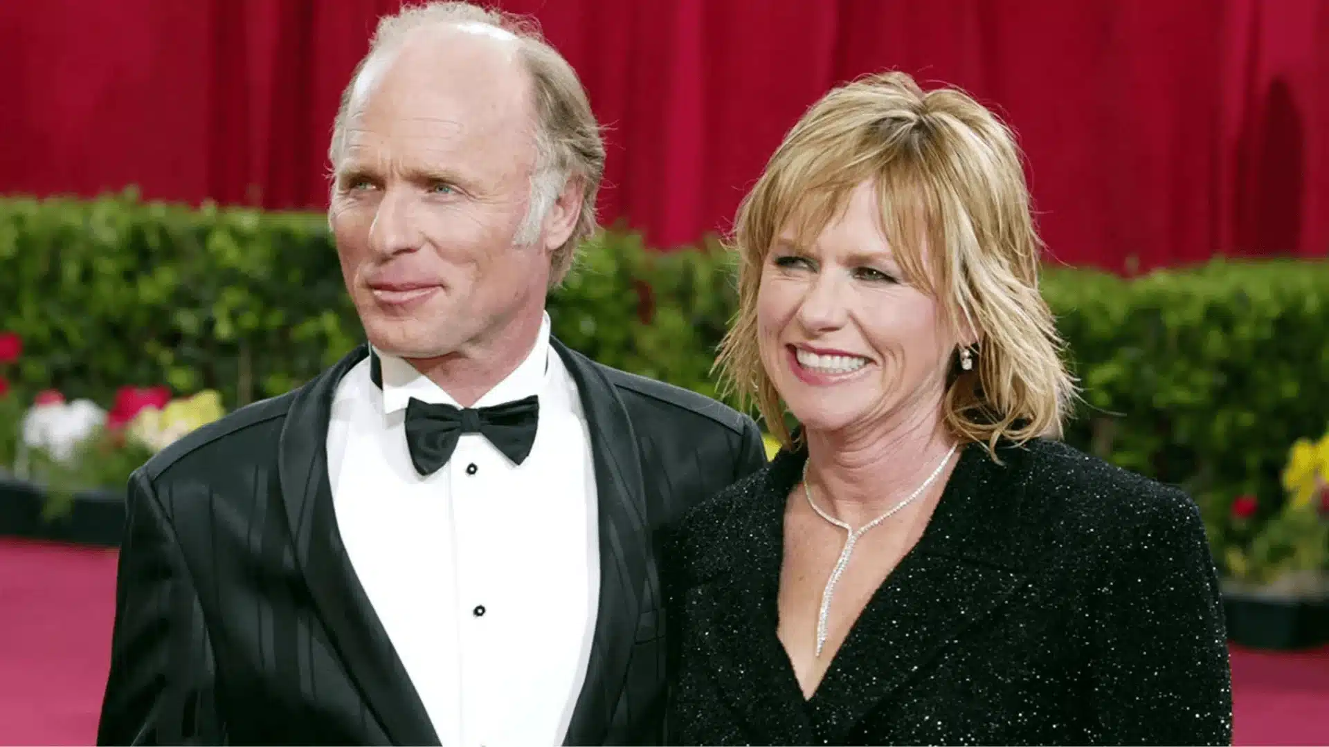 Ed Harris and Amy Madigan are posing together. He is wearing a formal tuxedo while she is smiling, wearing a textured black jacket