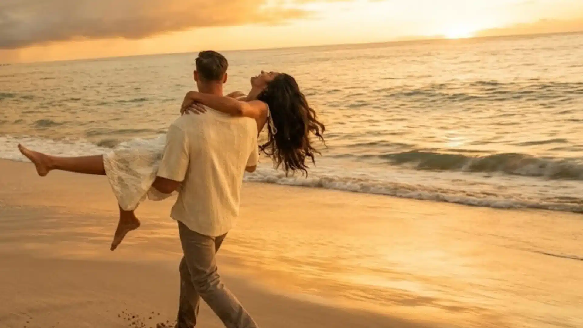 Man carries a woman in his arms along a sandy beach as the sun sets over the ocean waves with mountains in the distance