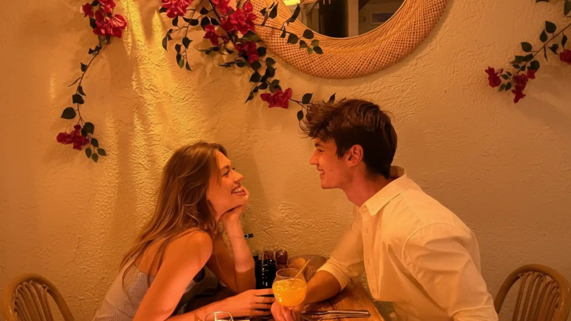 Smiling young couple looks at each other while sitting at a small table in a warm restaurant decorated with pink flowers