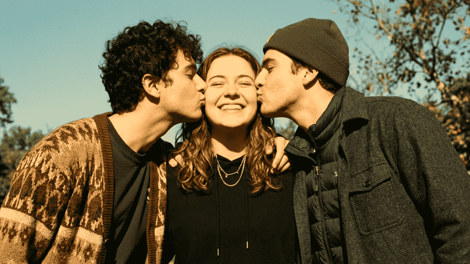 Two men kiss a smiling woman outdoors, showing a happy polyamorous relationship. (2)