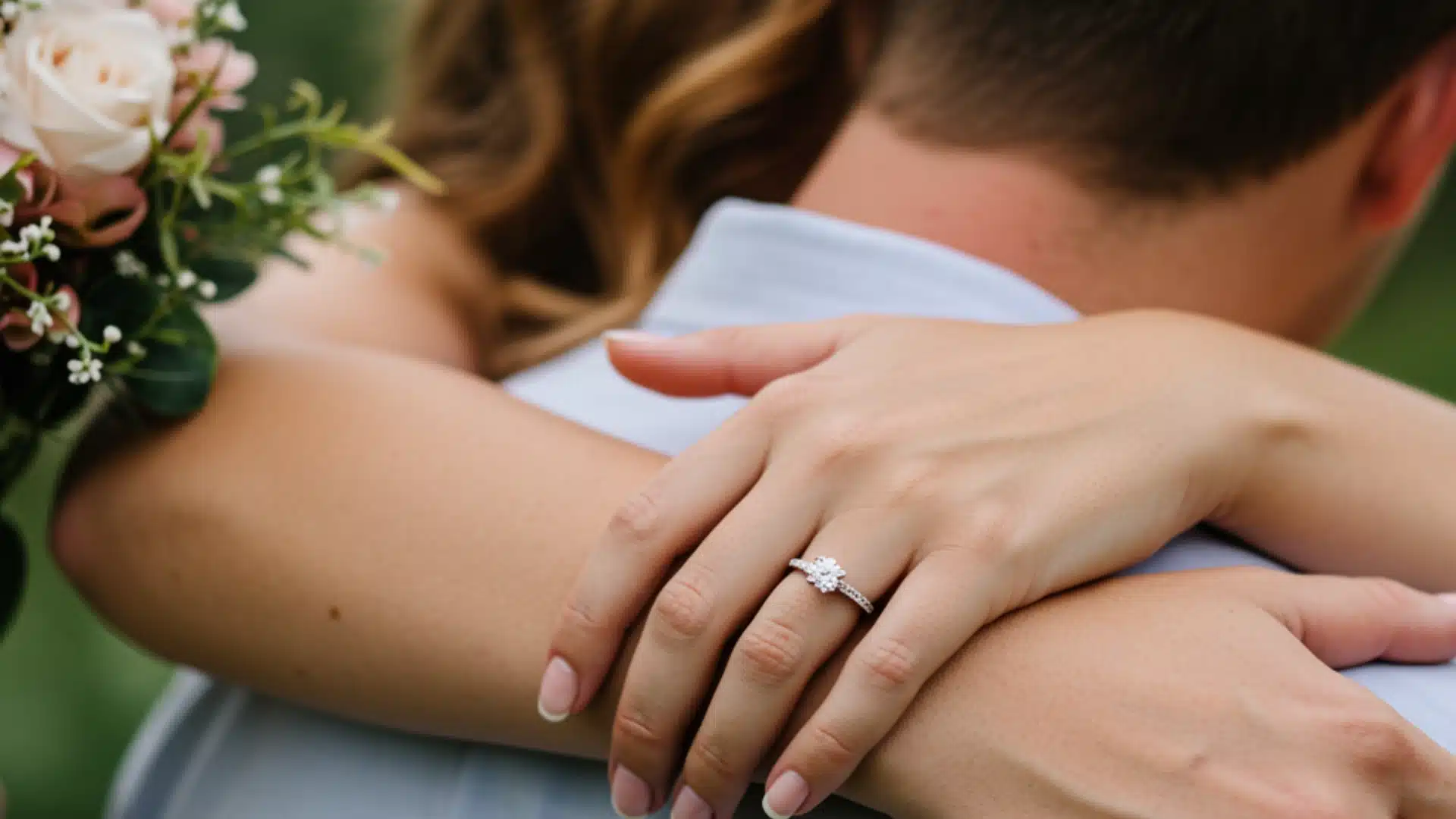 Close-up of a bride's hands with an engagement ring embracing a groom from behind, holding a small bouquet of pink and white roses.