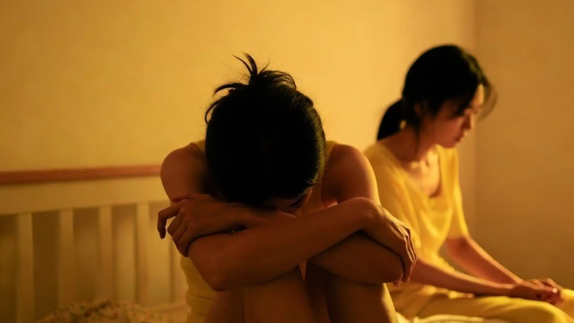 A lesbian couple sits on a bed in dim, warm lighting, one hunched over with her arms wrapped around her knees while the other looks away
