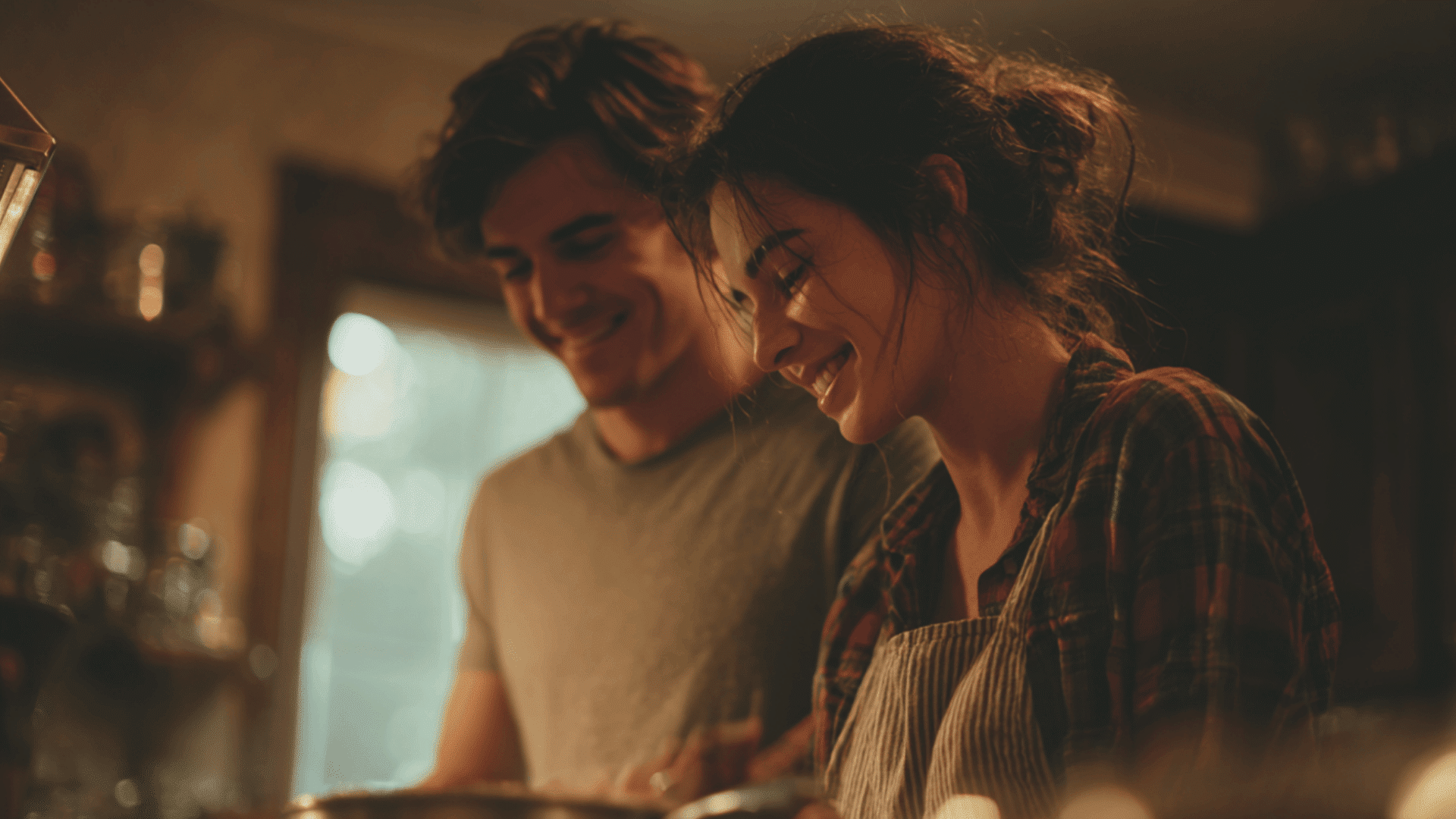 smiling couple laughing while cooking together in a kitchen with warm indoor lighting and the woman wearing a tan apron