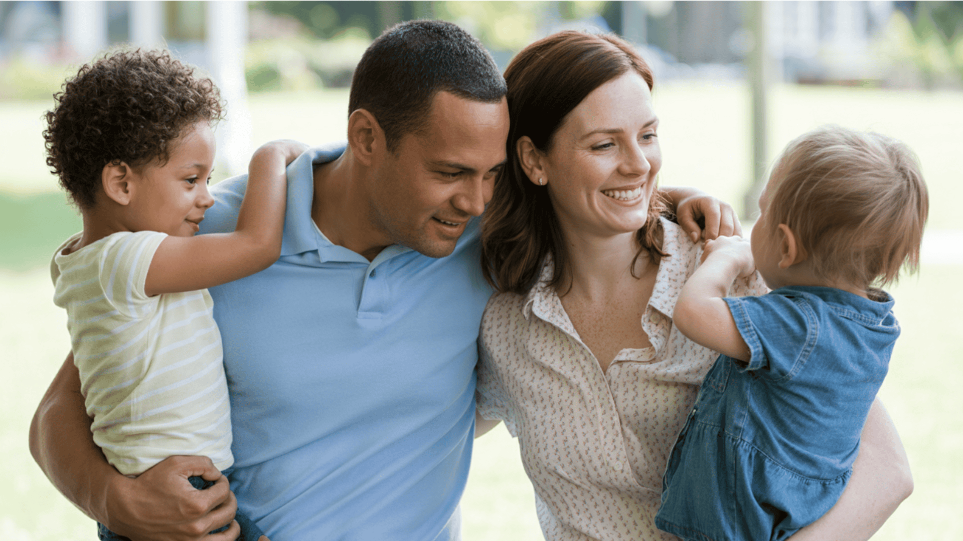 smiling parents each hold a young child while standing outside in a grassy park area on a clear and bright sunny day