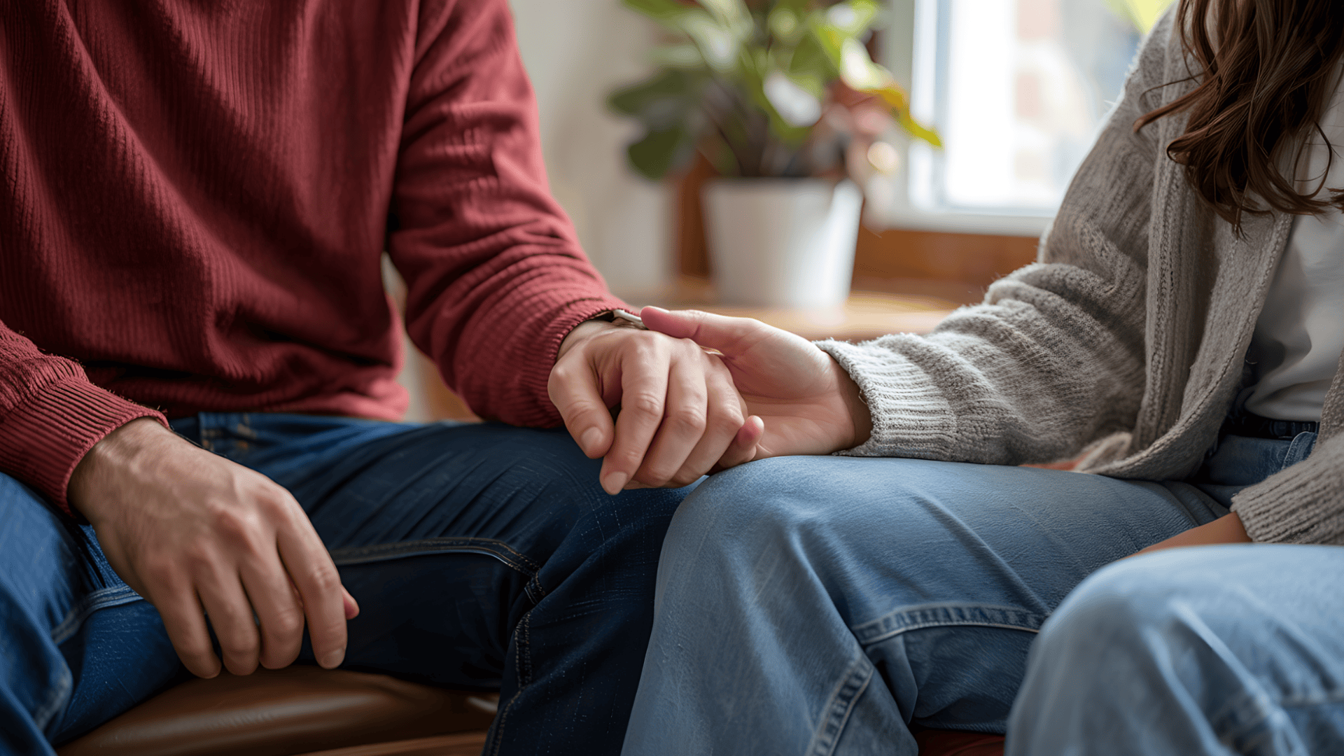 two people sitting side by side and holding hands in a gentle gesture of support with a small green plant in the background