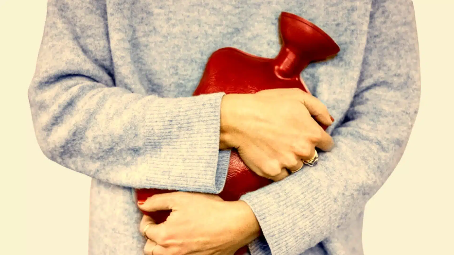 A woman in a soft blue sweater holding a red hot water bag against their lower abdomen, suggesting pelvic pain relief