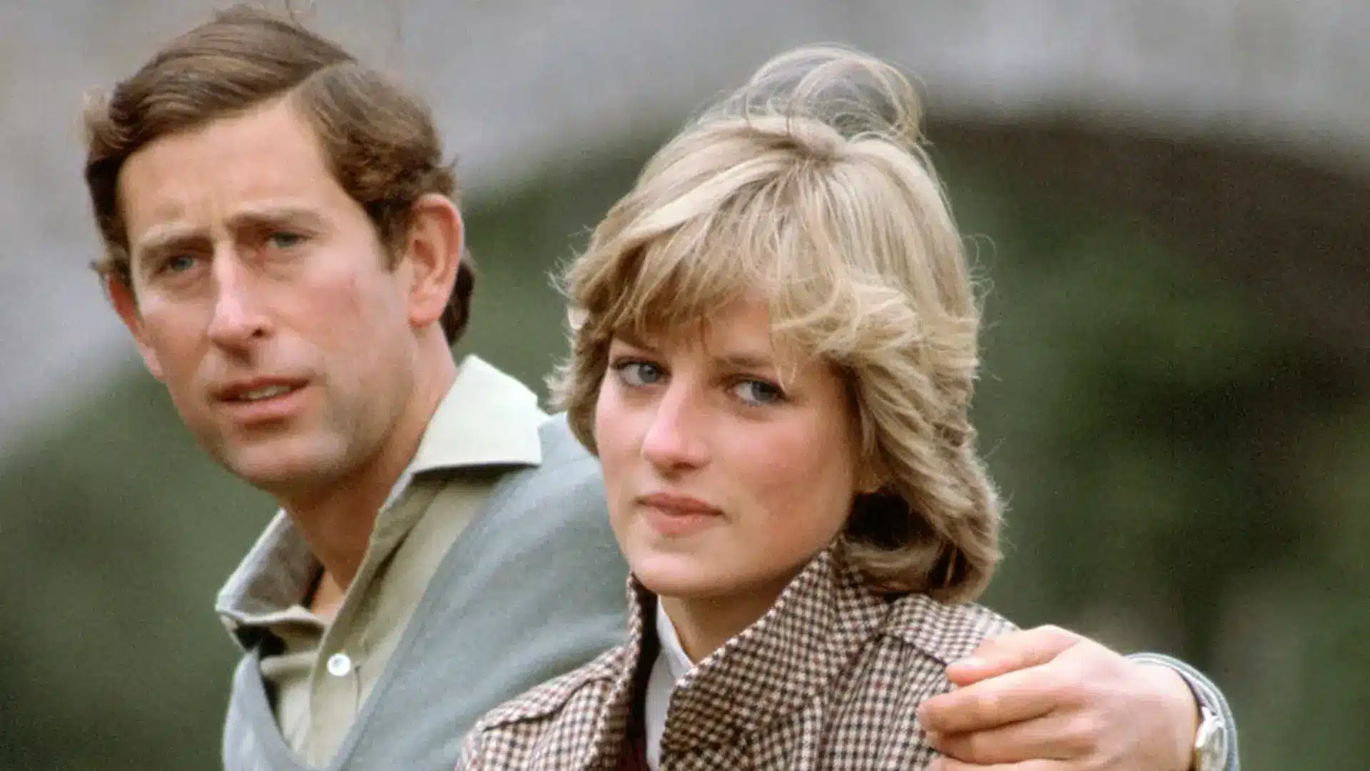 Prince Charles stands behind and has his hand on the shoulder of Princess Diana, both looking toward the camera outdoors