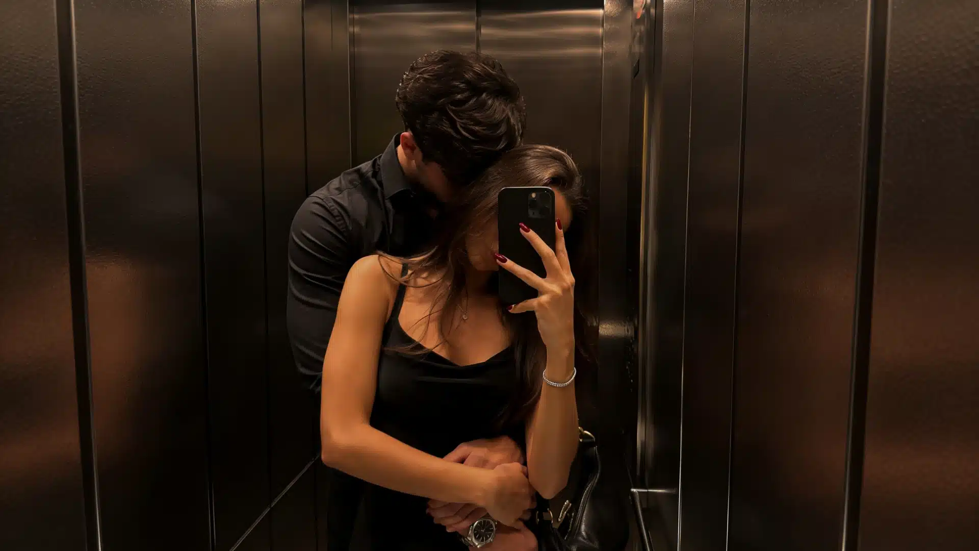 A couple takes a mirror selfie inside a dark elevator, embracing closely in black outfits