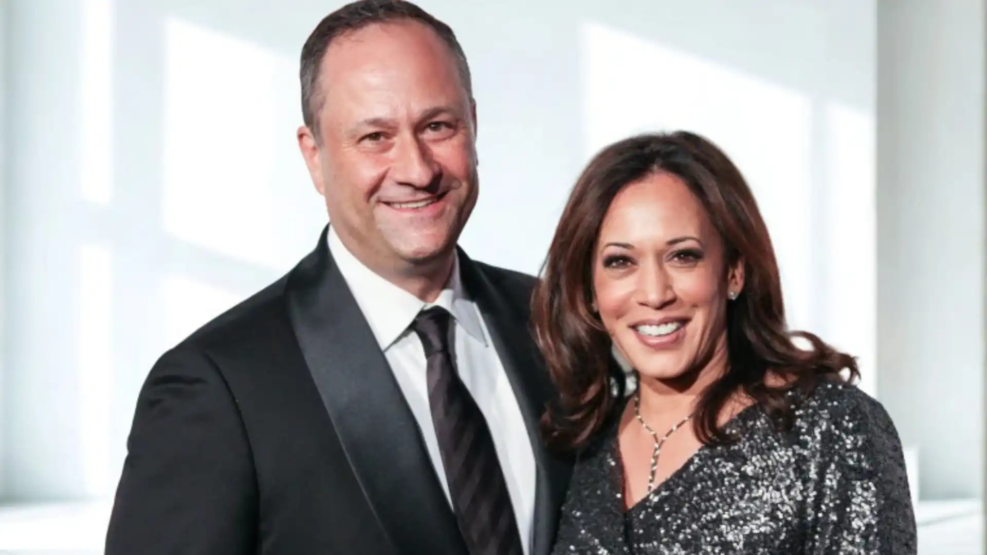 Kamala Harris and Doug Emhoff smile at the camera while dressed in formal attire against a bright, soft-focus background.