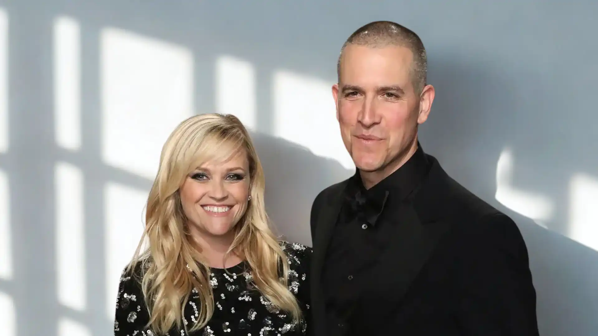 Reese Witherspoon and Jim Toth smile for a portrait against a light grey background featuring window shadows.