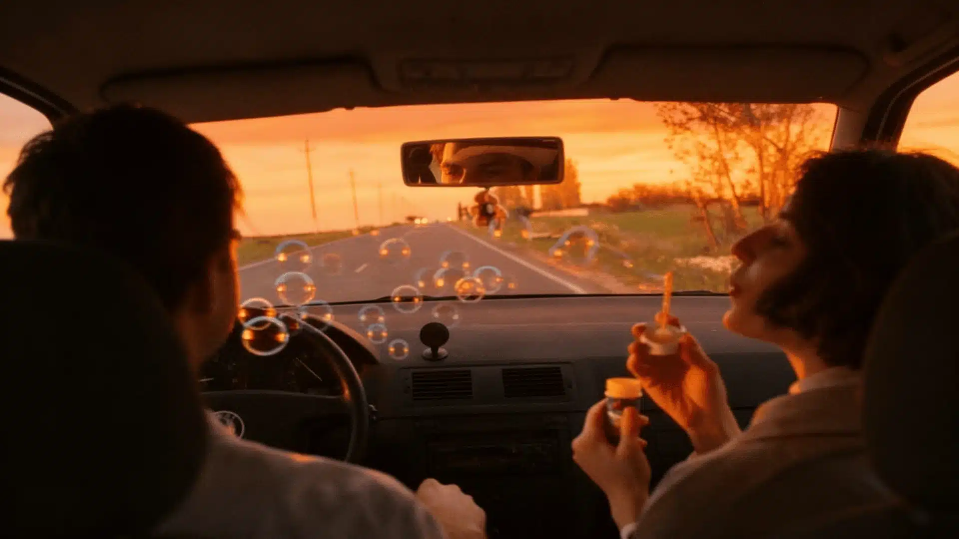Couple enjoying a sunset road trip date inside a car while blowing bubbles on empty highway