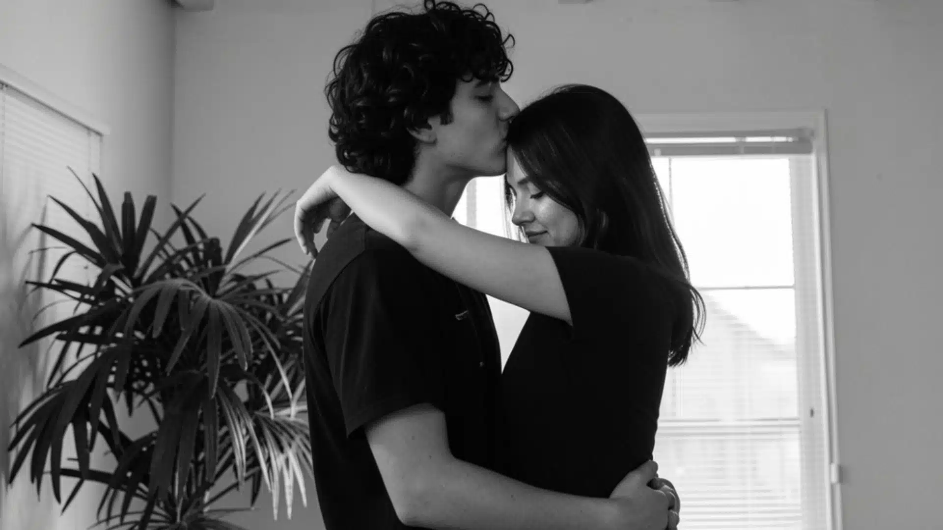Black and white photo of a young man kissing a young woman on the forehead while they embrace indoors next to a large houseplant and window.