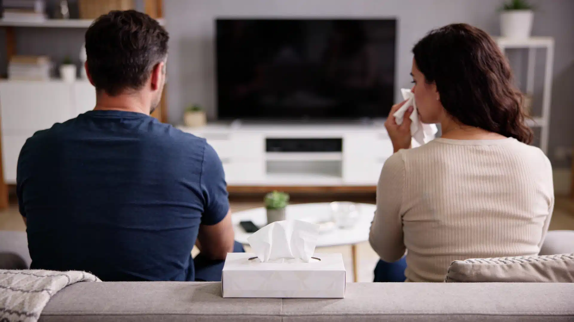 Man and woman sitting on a couch watching television, with the woman wiping her eyes with a tissue next to a tissue box