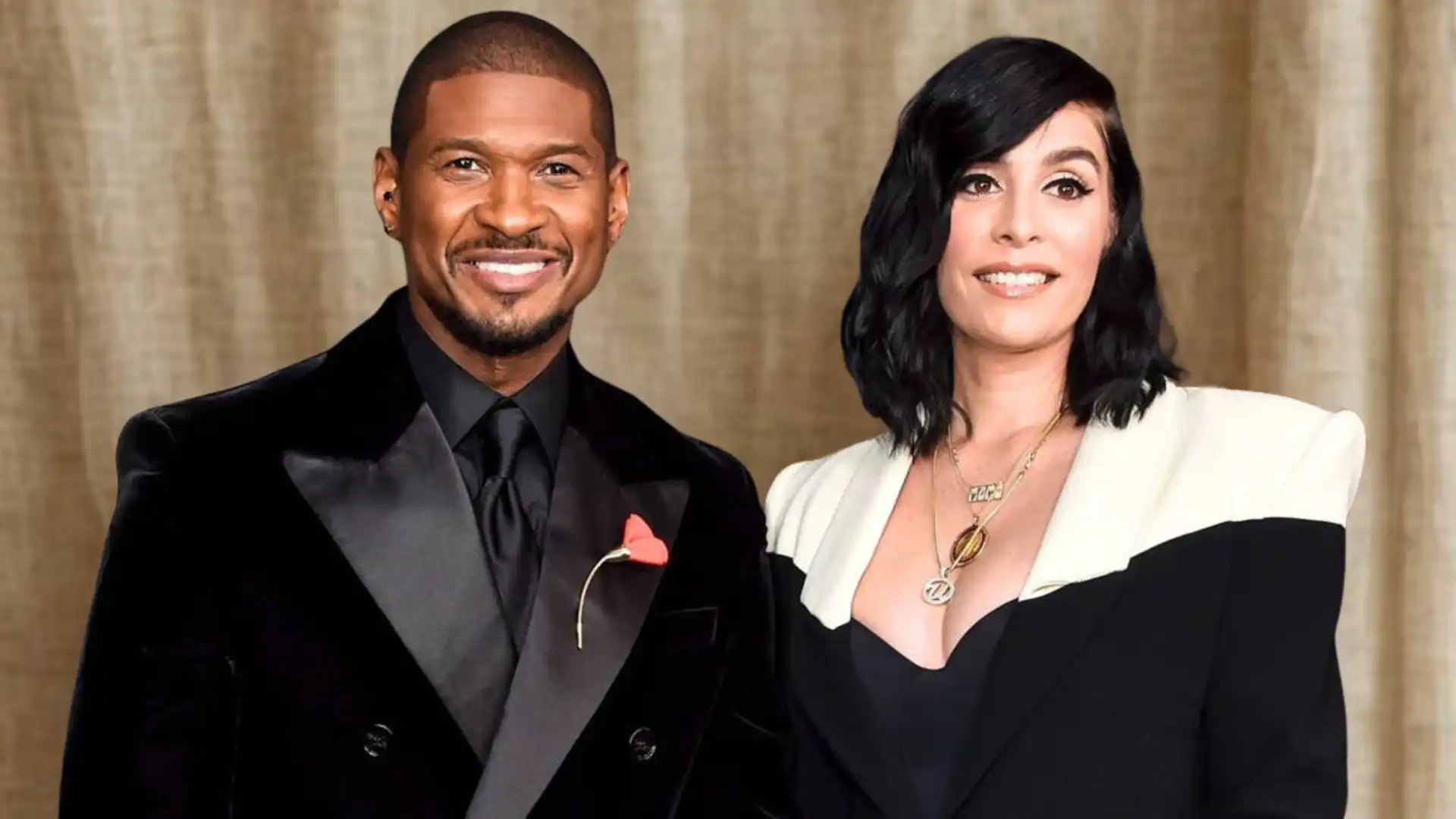 Usher smiles beside his partner, Jenn Goicoechea, who wears a black and white blazer, against a tan backdrop.