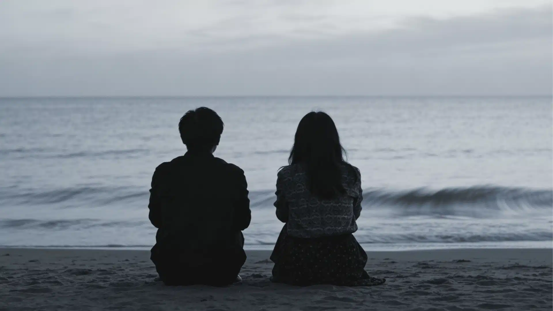Silhouette of a man and woman sitting side by side on a sandy beach, looking out at the ocean