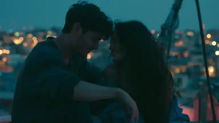 A young couple sits closely together on a rooftop at dusk, sharing an intimate moment with city lights blurred in the background.