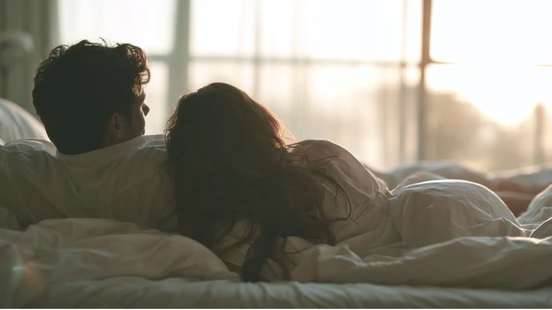 A couple sits together in bed beneath white blankets, gazing toward a sunlit window.