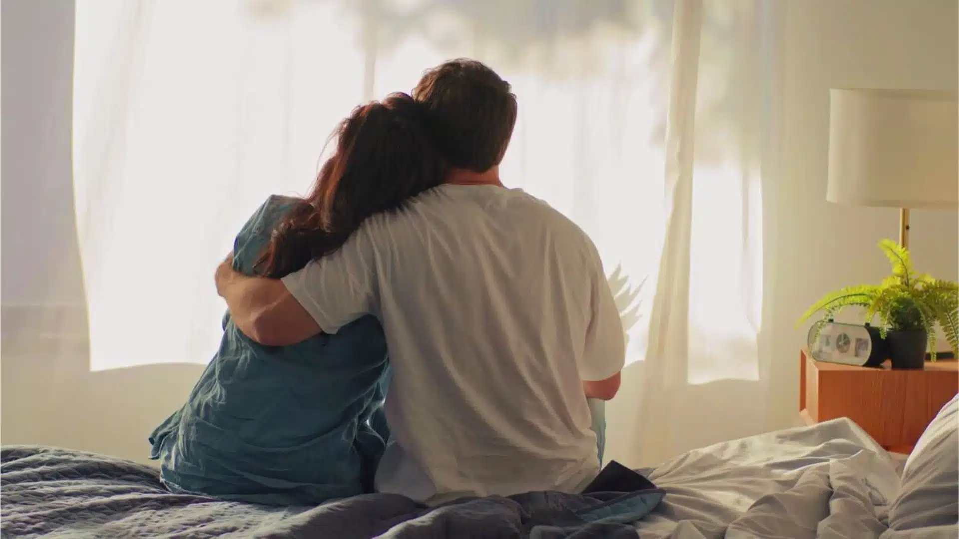 A man and woman sit together on a bed, facing a window with natural light streaming in.