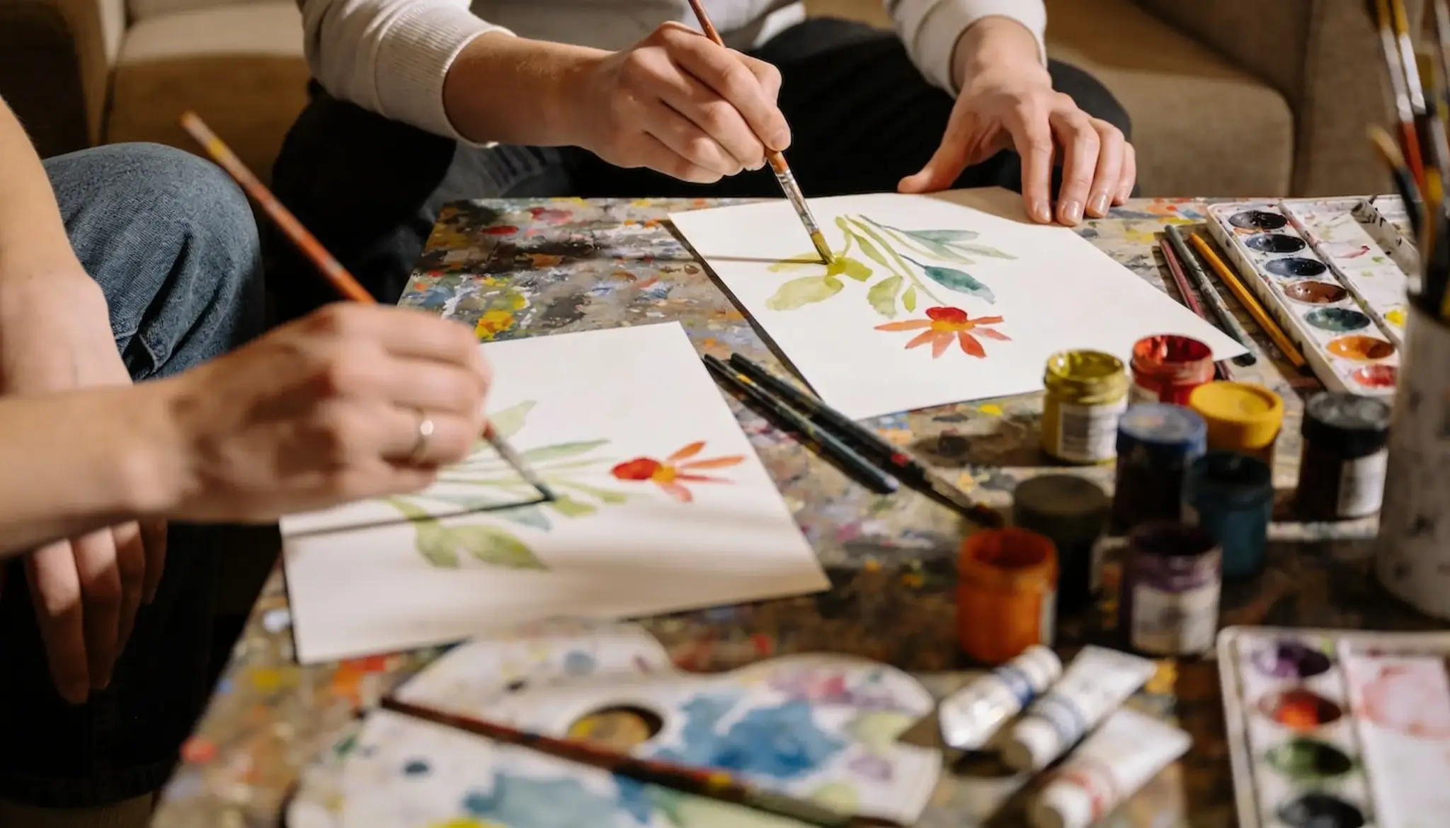 Two people are painting watercolor flowers on white paper with brushes and various paint supplies scattered on a colorful table.