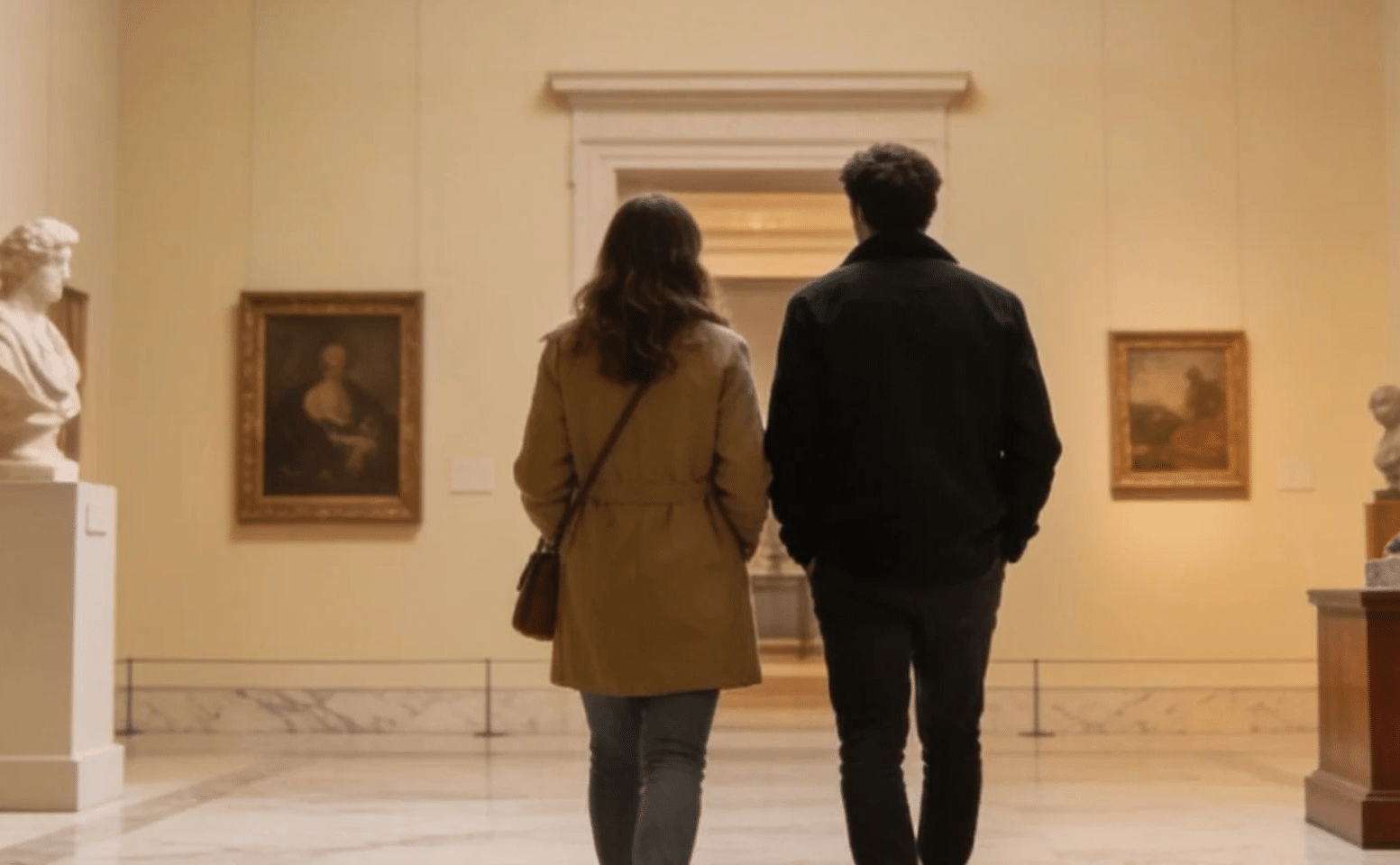 A man and woman walk together in a museum, engaged in conversation while observing the exhibits around them.