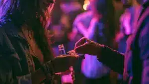 Close-up of hands exchanging a small folded packet in a dimly lit, neon-pink nightclub with blurred people in the background.