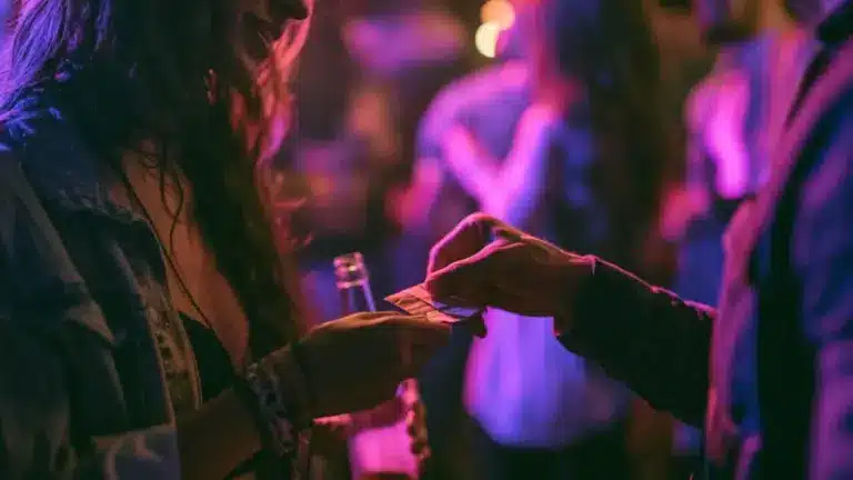 Close-up of hands exchanging a small folded packet in a dimly lit, neon-pink nightclub with blurred people in the background.