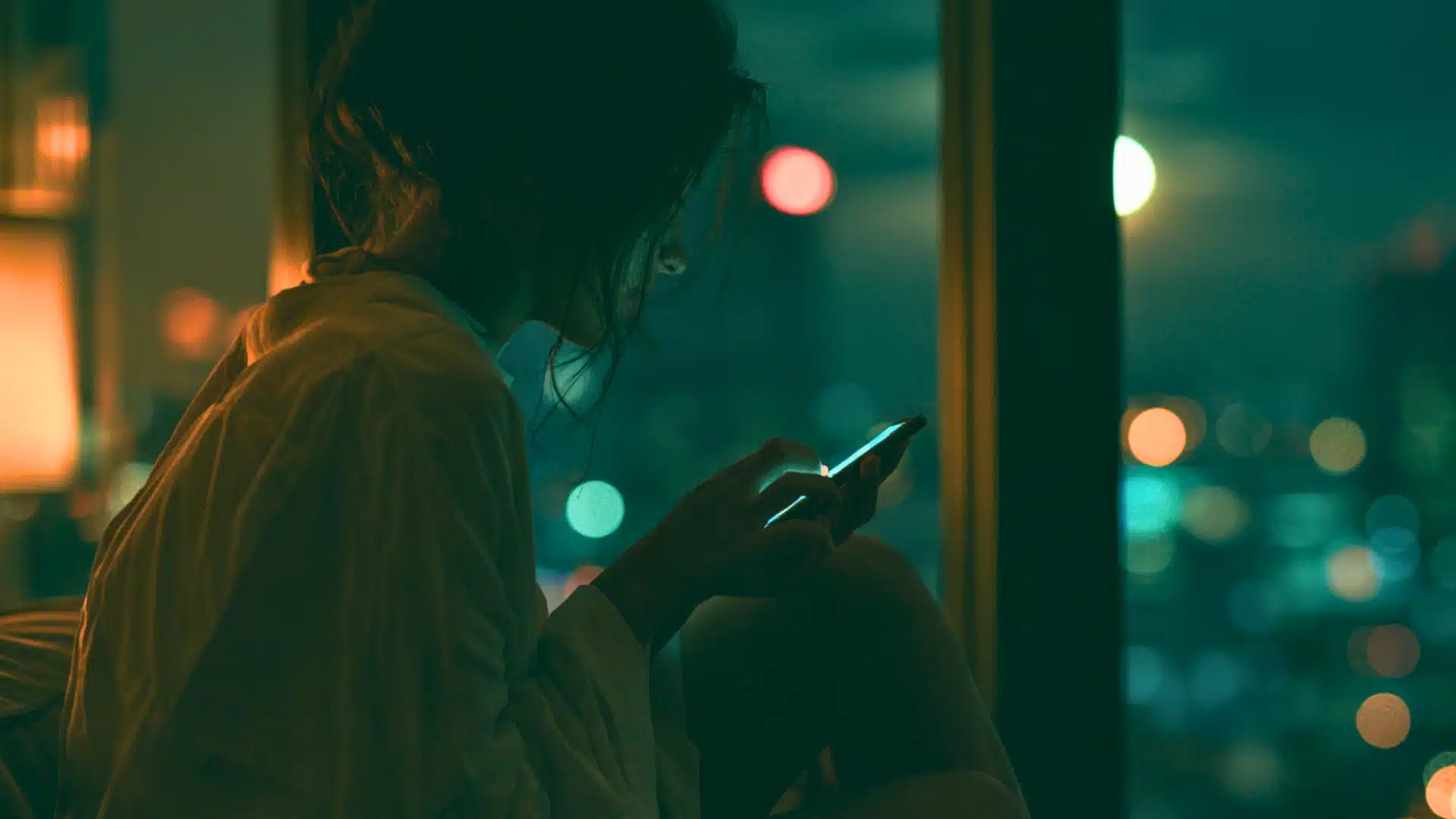 A person sitting by a window at night, illuminated by the screen of a smartphone, with a blurred cityscape visible in the background.
