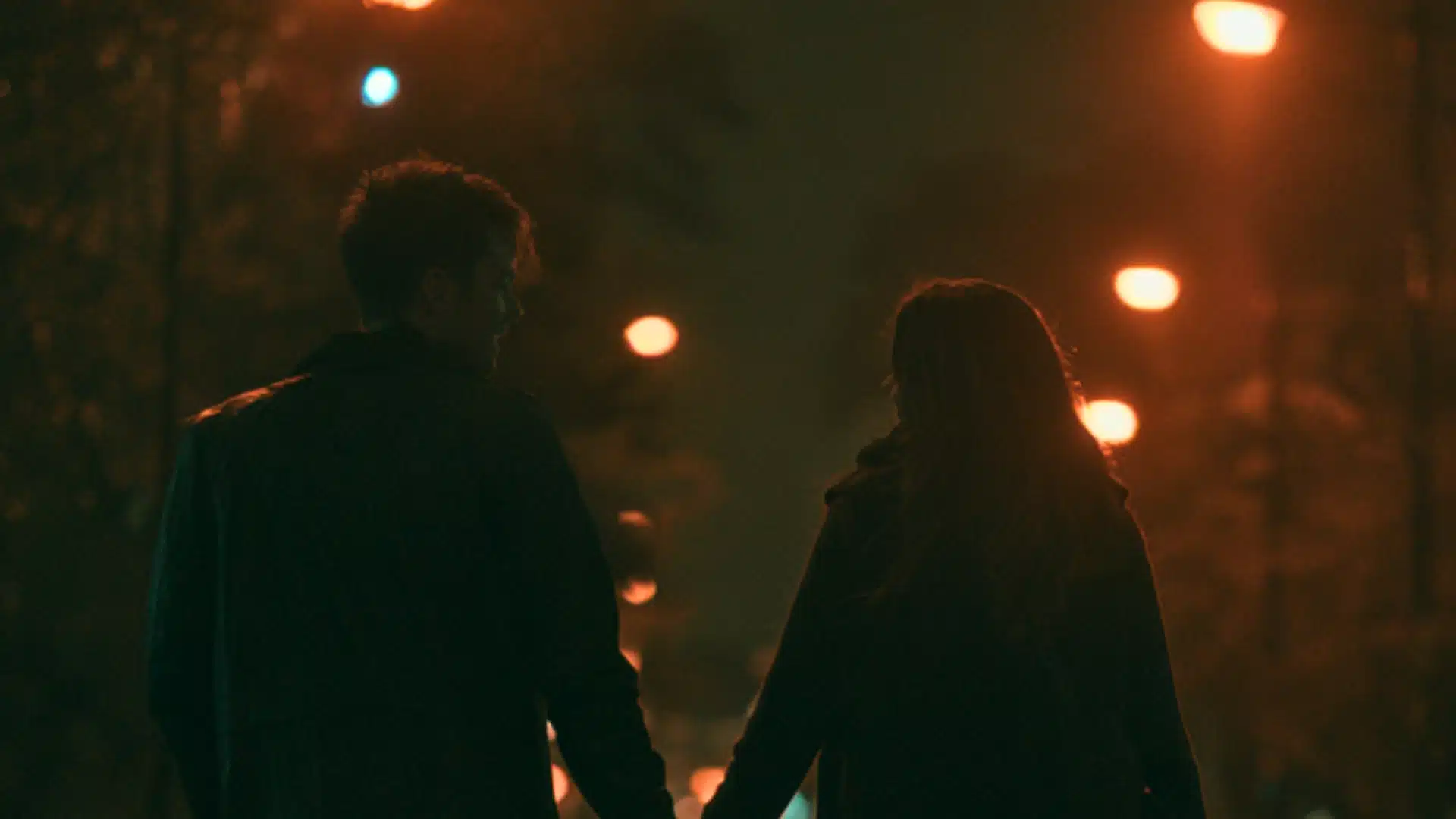 A young couple is walking hand in hand along a dimly lit street at night, illuminated by the warm, blurred glow of distant streetlights.